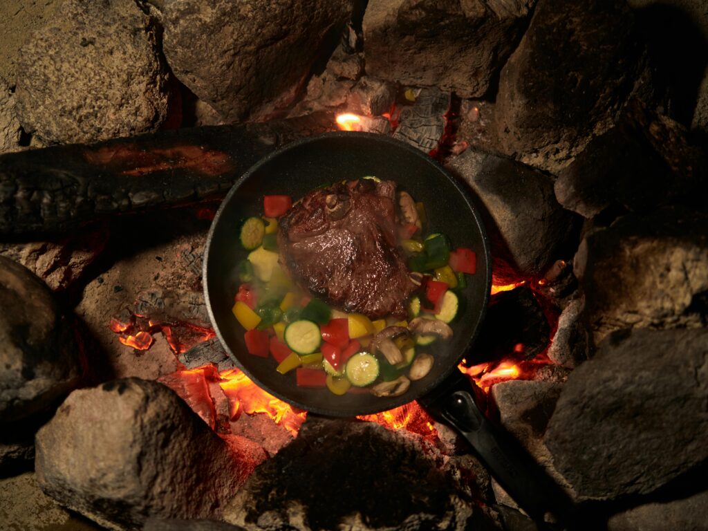 Sizzling beef steak with vegetables cooked over a campfire for an outdoor meal experience.