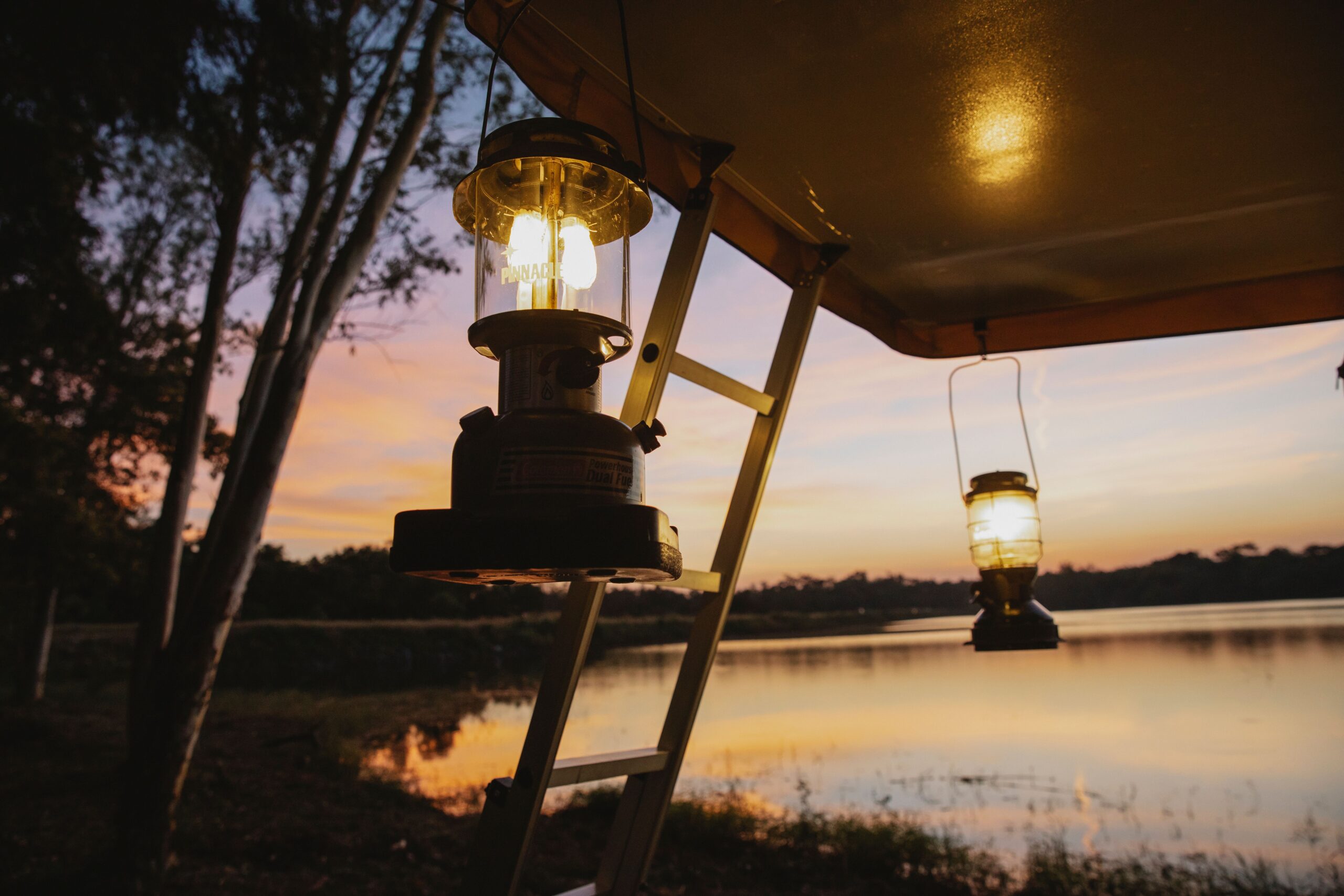 Camping pitfalls, what nobody tells you about your first camping trip Empty space under camping tent with lamps and stairways placed on coast of lake with thin trees in evening on sunset